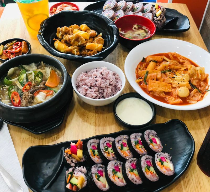 Ẩm thực Hàn Quốc bàn ăn Hàn Quốc tại Hanuri gồm tokbokki, canh bò hầm rau củ, kimbap gạo lứt, cơm tím, gà chiên phô mai và nhiều món ăn kèm, được bày trí hấp dẫn trên bàn gỗ.