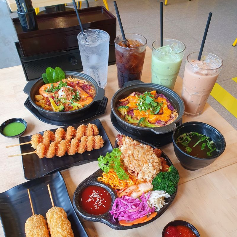 Set ăn Hàn Quốc tại Mì Cay Sasin gồm mì cay kimchi hải sản, cơm trộn, tokbokki, xiên chiên, súp kèm và 4 loại thức uống – bày trí hấp dẫn trên bàn gỗ trong không gian trẻ trung.