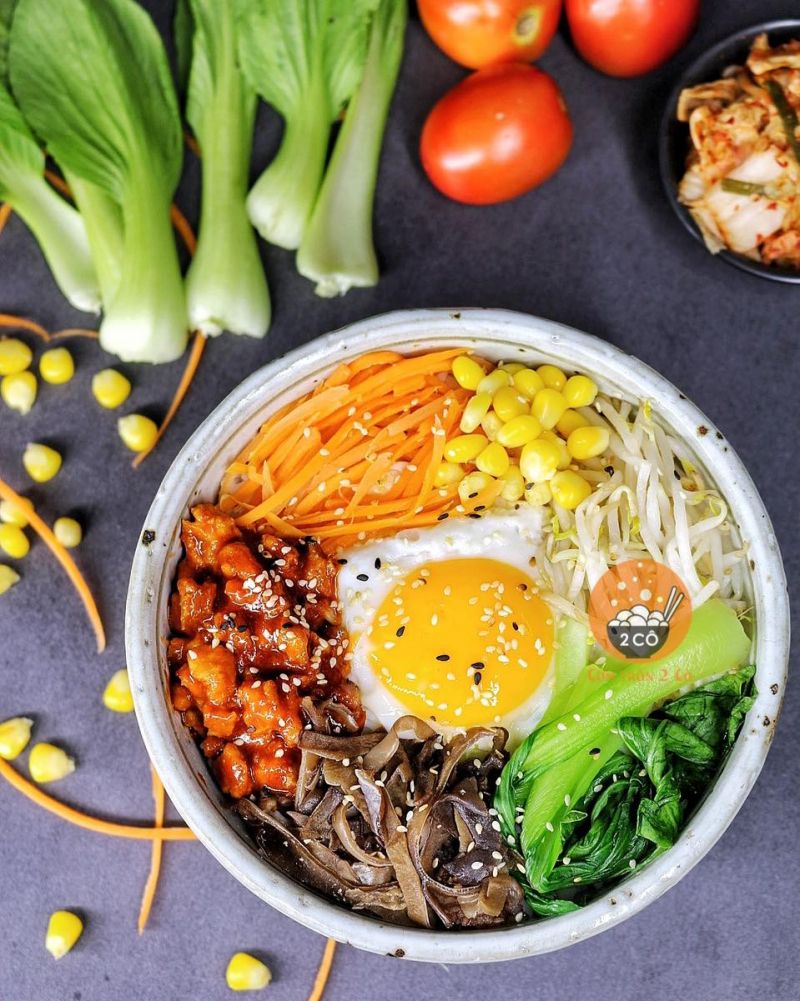 Cơm trộn ẩm thực Hàn Quốc với trứng ốp la, thịt sốt cay, nấm mèo, cải thìa, giá đỗ, cà rốt và bắp vàng – trang trí bắt mắt trong bát sứ trên nền bàn xám, kèm nguyên liệu tươi sống bên ngoài.