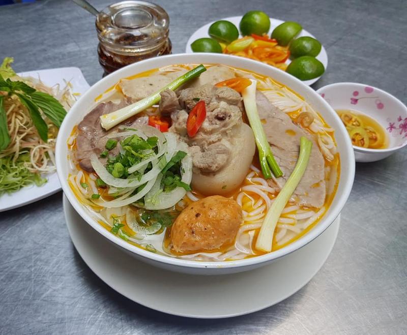 Tô bún bò Huế tại Bún Bò Hạnh gồm giò heo, chả, thịt bò lát, hành tây và hành lá, ăn kèm rau sống và nước chấm – đậm đà và đầy đặn.

