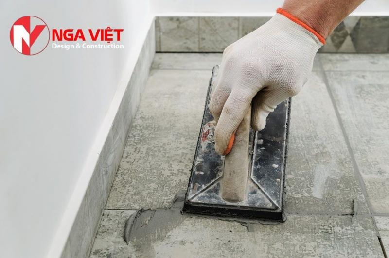Người thợ mang găng tay đang dùng bàn xoa xử lý lớp vữa chống thấm tại sàn nhà, logo Nga Việt ở góc trên cùng bên trái.