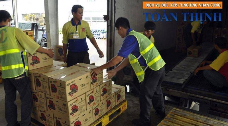 “Bốn công nhân Toàn Thịnh mặc áo vàng-xanh có phản quang đang bốc xếp thùng carton ‘Drinho’ lên pallet cạnh cửa container trong nhà kho.”