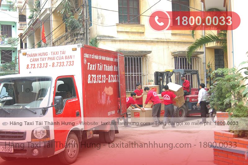 “Xe tải nhỏ màu đỏ ‘Taxi Tải Thành Hưng’ đậu trong sân hẻm; công nhân áo đỏ cùng xe nâng chất thùng carton, hotline 1800.0033 nổi bật, thể hiện dịch vụ vận chuyển 24/7.”