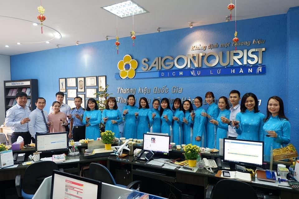 Nhân viên công ty du lịch Saigontourist mặc áo dài xanh đồng bộ, đứng thành hàng trong văn phòng, cùng nâng ly chúc mừng dưới logo thương hiệu nổi bật trên nền tường xanh.