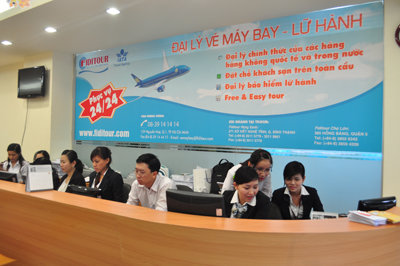 Nhân viên công ty du lịch Fiditour ngồi làm việc tại quầy tiếp tân, phía sau là bảng thông tin dịch vụ đặt vé máy bay và tour du lịch.