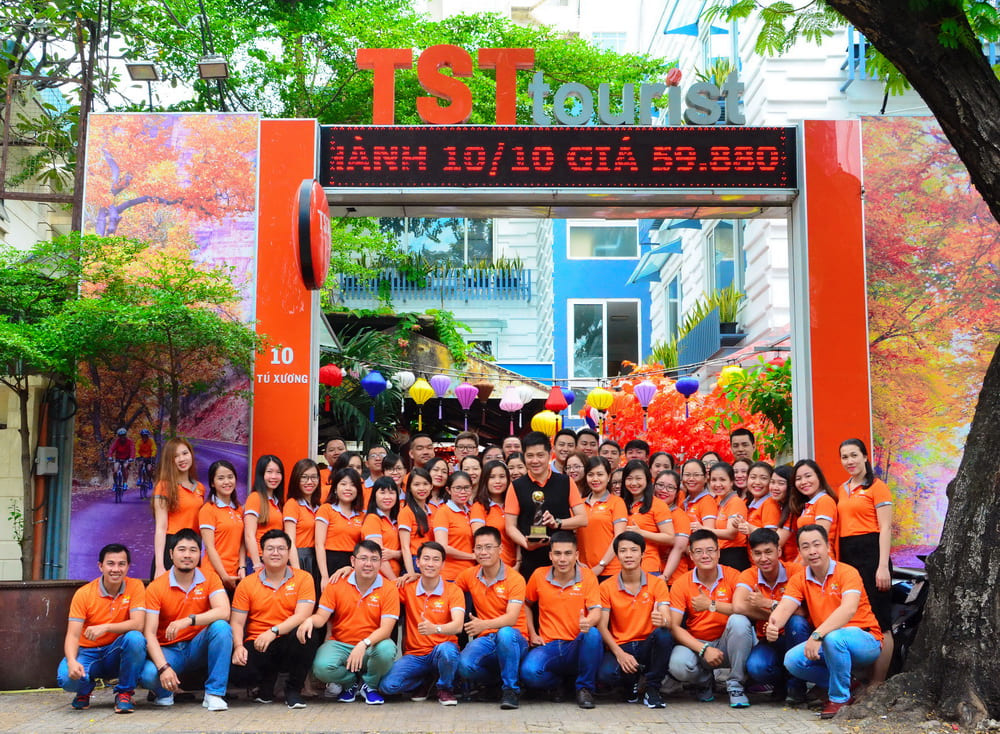 Tập thể nhân viên TST Tourist mặc áo cam chụp ảnh trước trụ sở chính tại TP.HCM, nổi bật với không gian rực rỡ đèn lồng và cây cảnh.