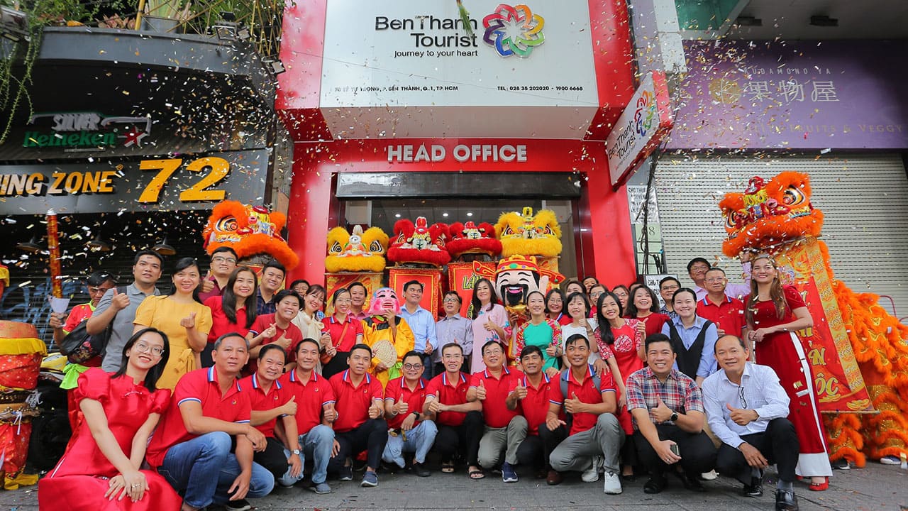 Tập thể nhân viên BenThanh Tourist chụp ảnh trước trụ sở chính trong lễ khai xuân, với múa lân, ông địa và không khí rộn ràng sắc đỏ.