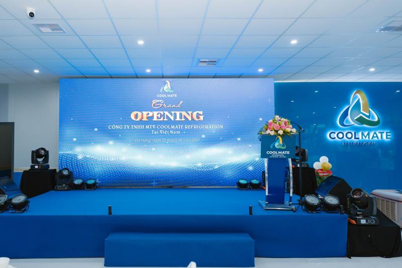 “Phòng hội trường với sân khấu xanh: màn hình LED rộng hiển thị dòng chữ ‘Grand Opening – Công ty TNHH MTV Coolmate Refrigeration’, bục phát biểu trang trí hoa, logo Coolmate phát sáng và dàn đèn moving head bố trí hai bên.”