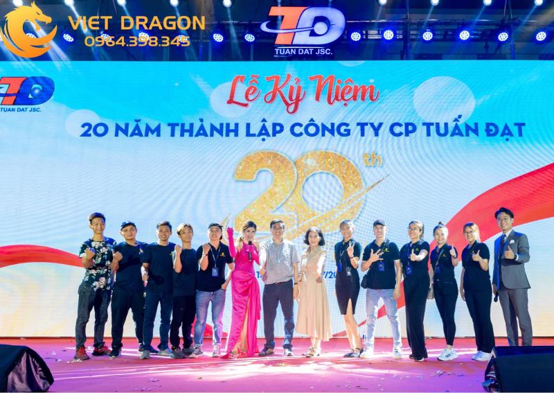 “Sân khấu sự kiện Viet Dragon Event: màn hình LED ghi ‘20 năm thành lập Công ty CP Tuấn Đạt’, người dẫn chương trình áo dài hồng đứng giữa dàn nhân viên và khách mời, ánh đèn xanh tím chiếu sáng phía trên.”
