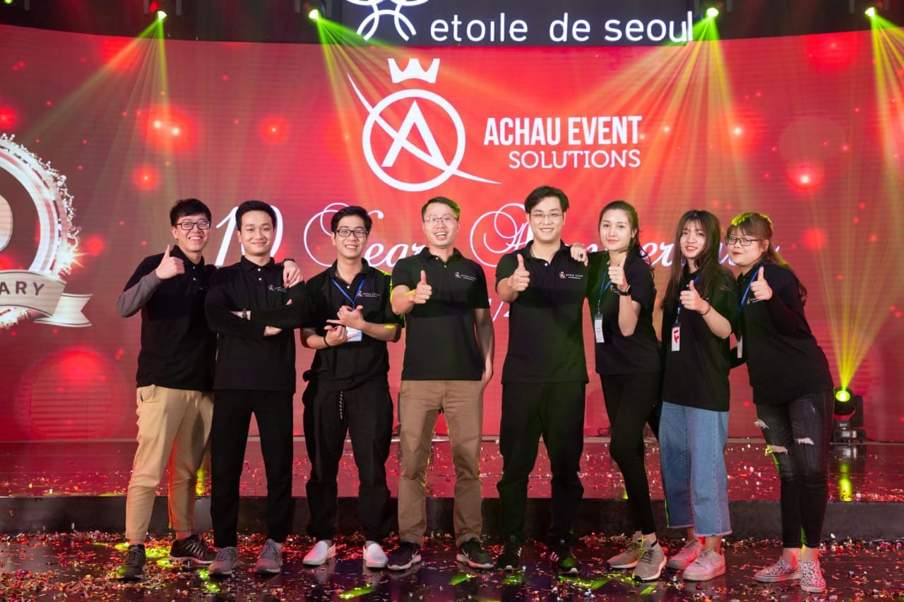 Đội ngũ nhân sự công ty tổ chức lễ kỷ niệm Á Châu Event Solutions tạo dáng trước sân khấu lễ kỷ niệm 10 năm thương hiệu với phông nền đỏ và logo nổi bật.