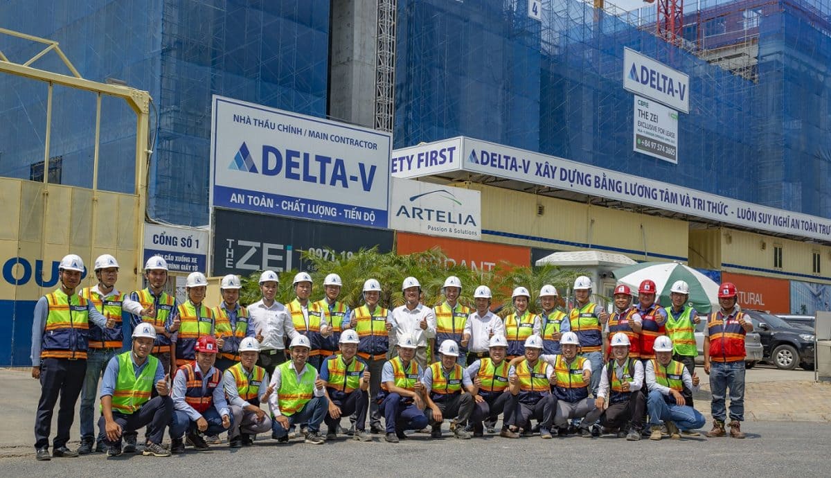 Nhóm kỹ sư và công nhân công ty xây dựng Delta chụp ảnh tại công trình đang thi công, với khẩu hiệu “An toàn – Chất lượng – Tiến độ” nổi bật phía sau.