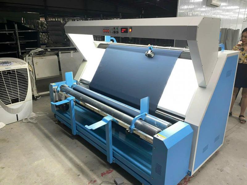 Máy kiểm vải tự động màu xanh, tích hợp đèn chiếu sáng và trục cuộn vải trong xưởng may TP.HCM.