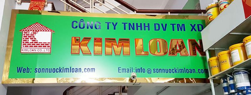 “Bảng hiệu xanh chữ vàng ‘Kim Loan’ treo trên cầu thang, kệ bên phải xếp thùng sơn nhiều màu; không gian bên trong đại lý sơn gốc nước VOC thấp.”

