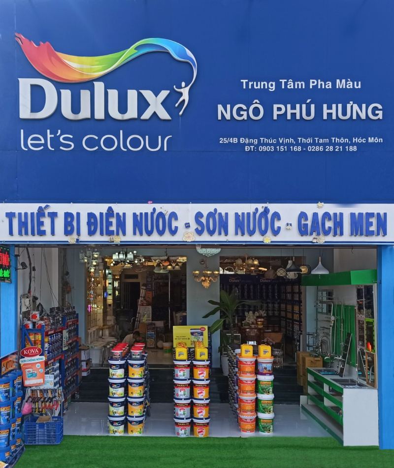 “Cửa hàng Dulux Ngô Phú Hưng tại 25/4B Đặng Thúc Vịnh, Hóc Môn: biển hiệu xanh ‘Dulux let’s colour’, dãy thùng sơn đủ màu xếp trước lối vào, bảng ‘THIẾT BỊ ĐIỆN NƯỚC – SƠN NƯỚC – GẠCH MEN’ phía trên.”