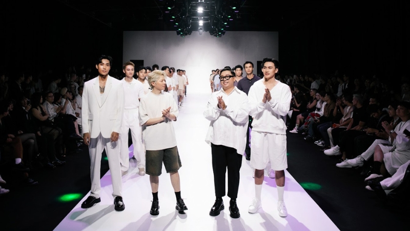 Dàn người mẫu nam trẻ trung vẫy chào khán giả cuối show runway, ánh đèn spotlight chiếu dọc sàn diễn trắng.