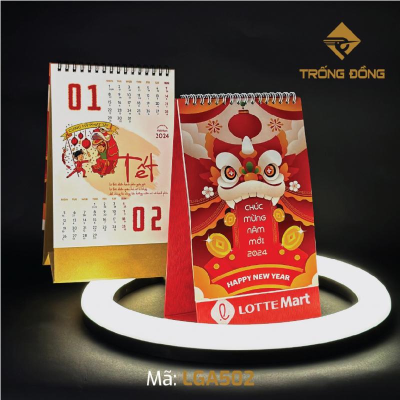 Hai cuốn lịch bàn Tết 2024 của In Trống Đồng, bìa đầu lân đỏ vàng và trang lịch tháng 1–2 với họa tiết múa lân, đặt trên vòng đèn sáng.