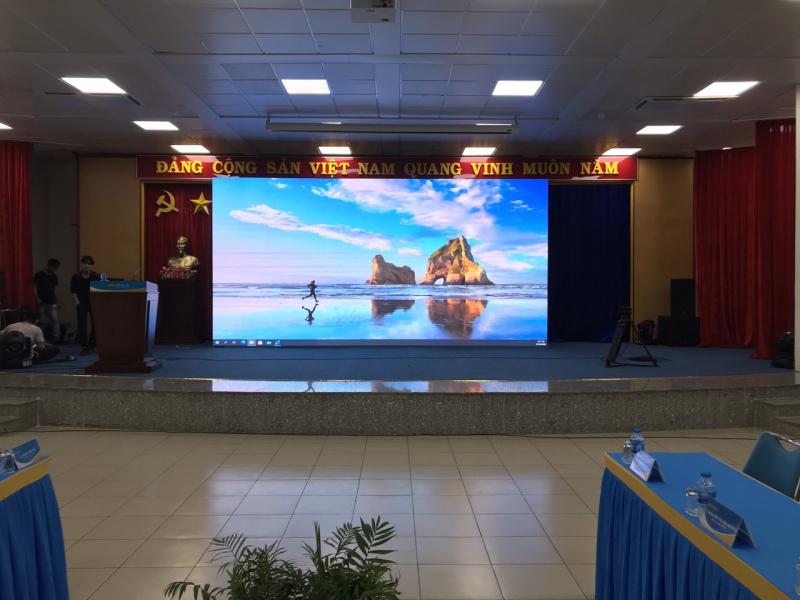 “Hội trường với bục phát biểu, tượng Bác Hồ, biểu ngữ ‘Đảng Cộng Sản Việt Nam Quang Vinh Muôn Năm’ và màn hình LED Việt Tin khổ lớn chiếu hình bãi biển, minh hoạ giải pháp LED Nationstar điều khiển cloud.”