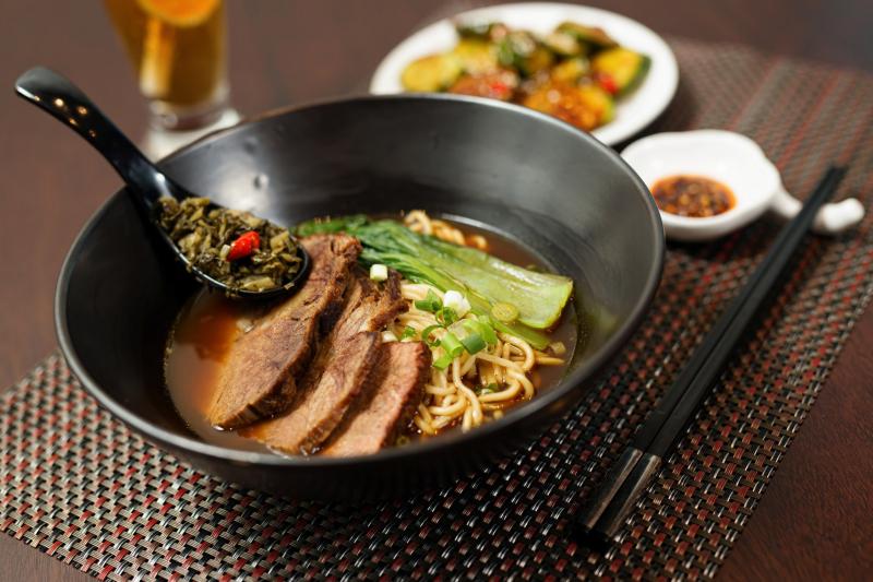 Tô mì bò Đài Loan tại Cow Mee Inn với thịt bò lát mỏng, rau cải xanh, cải muối và nước dùng đậm đà, đặt trên bàn ăn sang trọng cùng đũa và thìa đen.