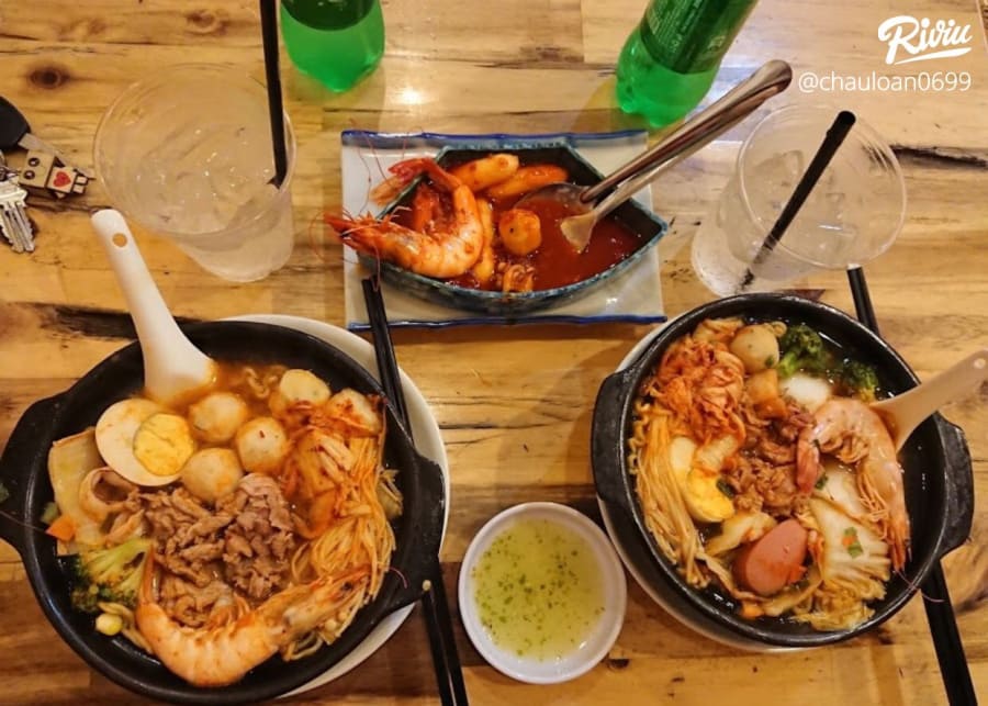 Hai tô mì cay thập cẩm cùng tokbokki và nước chấm được bày trên bàn gỗ tại một quán mì cay phong cách Hàn Quốc.