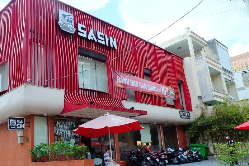 Quán mì cay Sasin màu đỏ với bảng hiệu lớn và khách xếp xe máy bên ngoài, chuyên phục vụ mì cay 7 cấp độ Hàn Quốc.