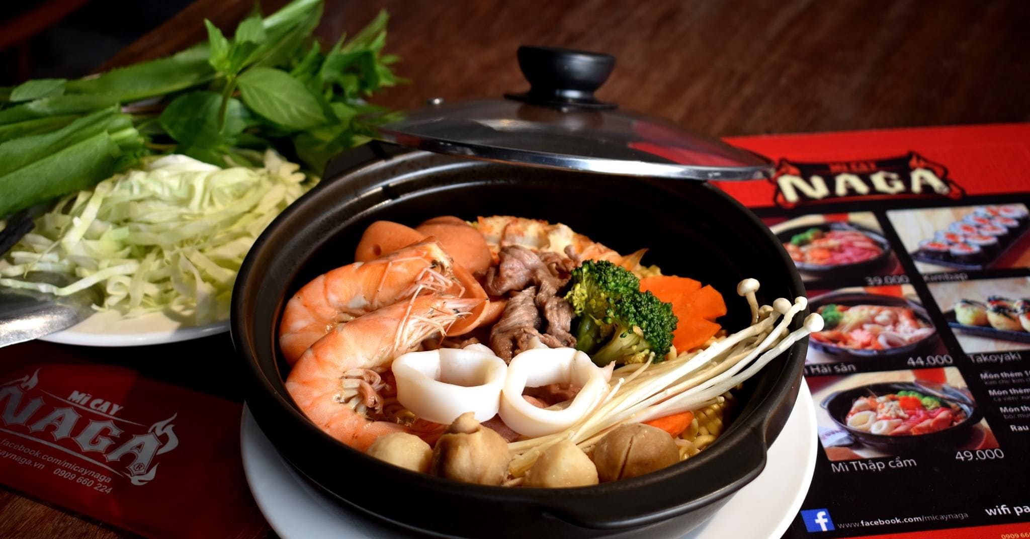Tô mì cay hải sản tại quán mì cay Naga với tôm, mực, thịt bò, rau củ, đặt cạnh menu và rau sống.