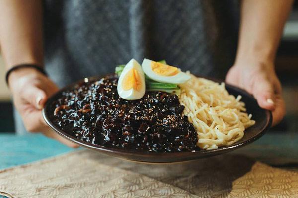 Phần mì tương đen Hancook gồm mì trụng vàng dai, sốt đậu đen bóng mịn kèm trứng luộc và dưa leo, được bày trí trên đĩa trước khi đóng hộp – lựa chọn lý tưởng cho dân văn phòng và học sinh.