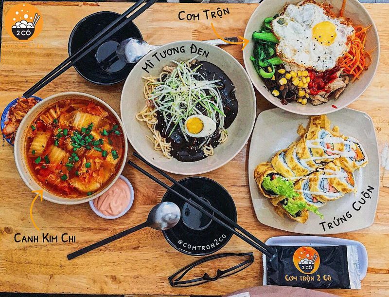 Ảnh chụp bàn ăn tại quán Cơm Trộn 2 Cô với mì tương đen kèm trứng lòng đào, cơm trộn rau củ, canh kimchi và trứng cuộn kiểu Hàn, bày trí gọn gàng và hấp dẫn.