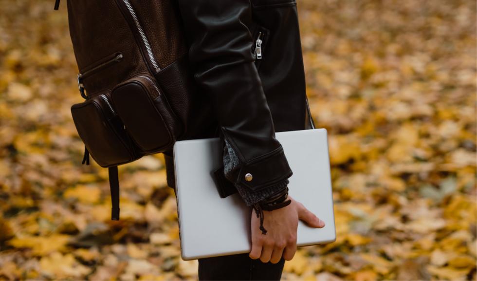 Nam sinh viên cầm laptop giá rẻ và balo da đi bộ trên thảm lá mùa thu