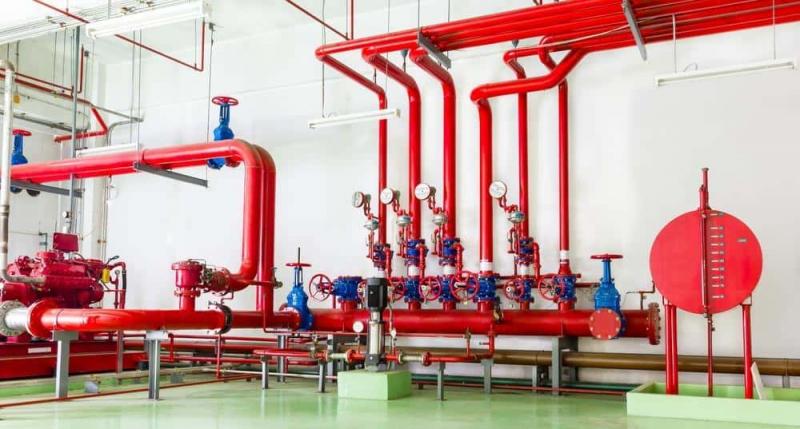 Dàn ống chữa cháy sơn đỏ với nhiều van, đồng hồ áp suất và ống góp ngang bên trong phòng kỹ thuật phòng cháy chữa cháy, sàn epoxy xanh.