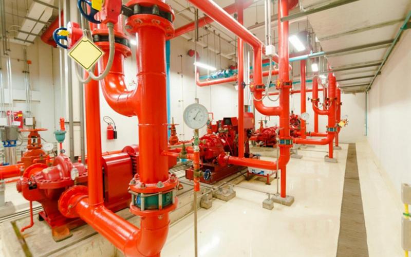 Hệ thống ống thép sơn đỏ nối các bơm chữa cháy trong phòng kỹ thuật PCCC, kèm đồng hồ áp suất và van điều khiển.