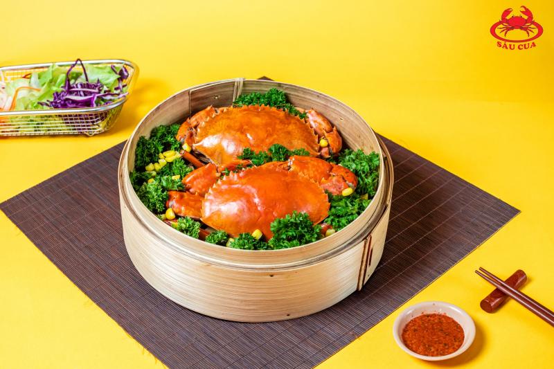 Hai con cua đỏ óng, đặt trong xửng tre hấp cùng rau xanh, bày trên bàn với nền vàng rực – món đặc trưng tại quán Út Cà Mau chuẩn vị miền Tây.