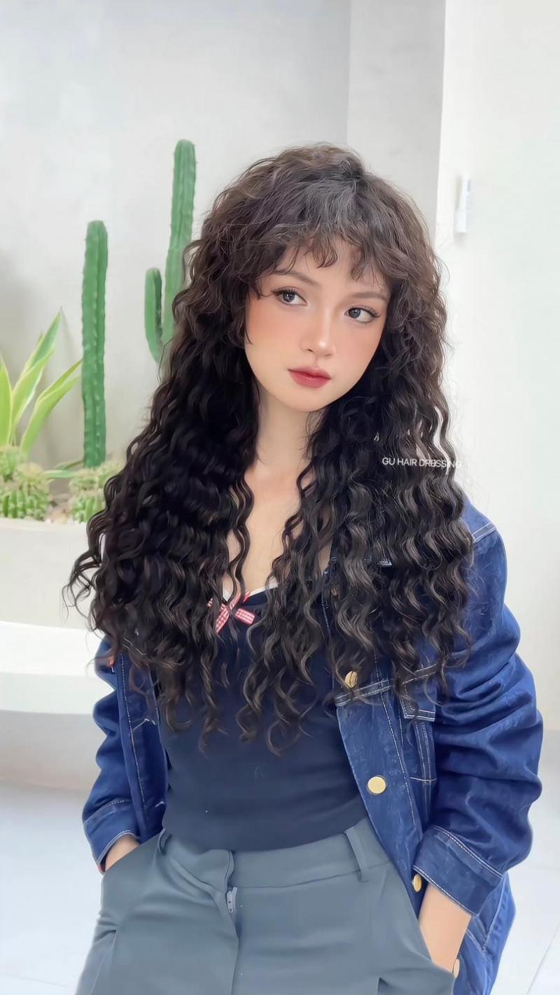 Cô gái với mái tóc uốn xoăn hippie dài, bồng bềnh, mặc áo khoác denim xanh, tạo dáng trước nền trắng với cây xương rồng phía sau – kiểu tóc được thực hiện tại Gu Hair Salon TP.HCM.

