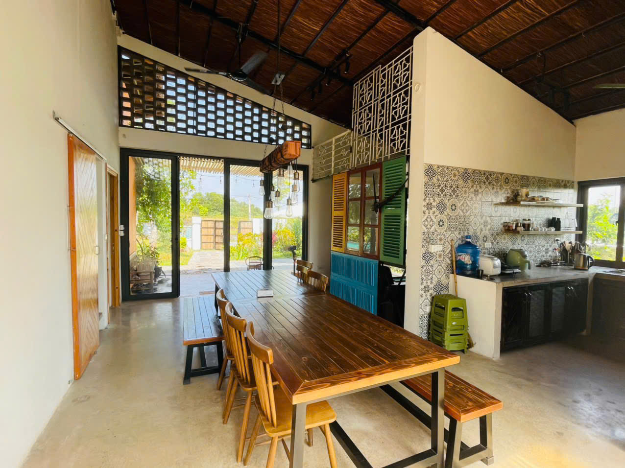 Không gian phòng ăn và bếp của một homestay thiết kế mộc mạc với bàn gỗ, trần mây tre và cửa kính hướng ra sân vườn.